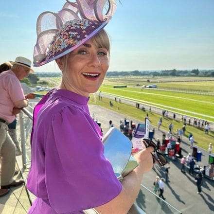 An attractive lady wearing a purple dress and floral fascinator smiling as she stands on a balcony overlooking Wetherby Racecourse on Ladies' Evening